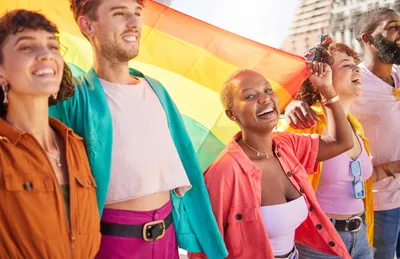 Four smiling young people in colorful outfits, waving a large Pride flag.