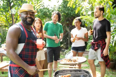 Sweaty man barbecues while holding a drink, with his guests looking at each other with concerned looks on their faces.